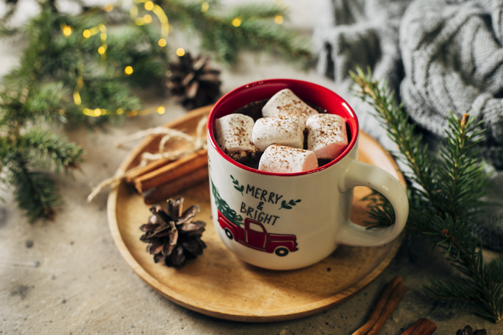 Hot chocolate with marshmallows and candy cane in holiday mug. Fir tree branch, cozy gray sweater on background. Cozy seasonal Christmas holidays concept